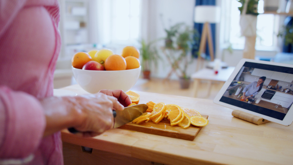 Attractive senior woman preparing food in kitchen indoors, following food vlogger on tablet.