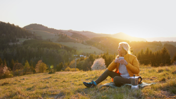 An attractive senior woman sitting outdoors in nature at sunset, relaxing with coffee.