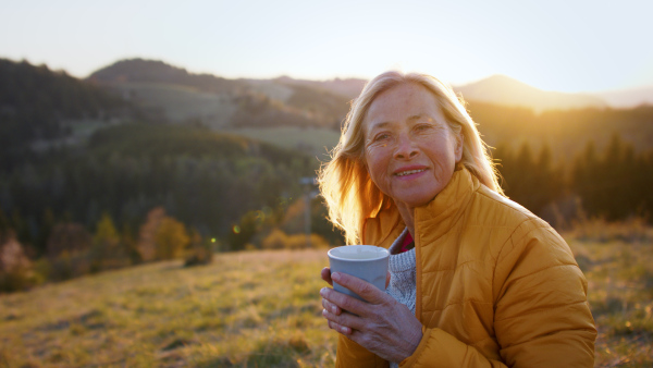 Attractive senior woman outdoors in nature at sunset, holding cup and looking at camera.