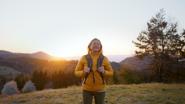 Attractive senior woman with backpack walking outdoors in nature at sunset, hiking.