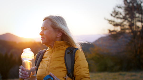 Attractive senior woman hiker walking outdoors in nature at sunset, drinking water.