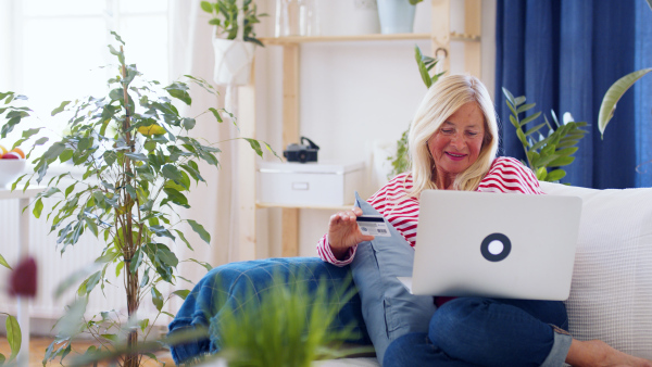 Attractive senior woman with laptop and credit card sitting indoors on sofa, online payment concept.