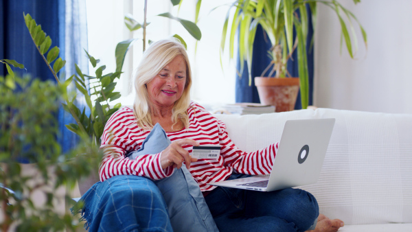 Attractive senior woman with laptop and credit card sitting indoors on sofa, online payment concept.