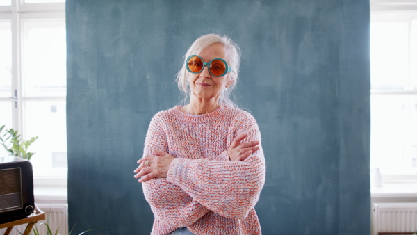 Portrait of senior woman with eccentric sunglasses standing indoors against dark background, looking at camera.