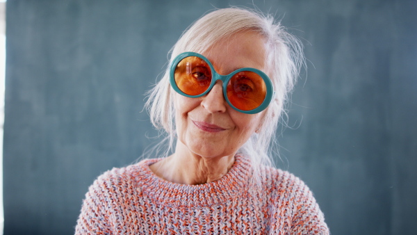 Portrait of senior woman with eccentric sunglasses standing indoors against dark background, looking at camera.