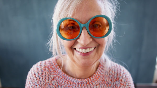 Portrait of senior woman with eccentric sunglasses standing indoors against dark background, looking at camera.