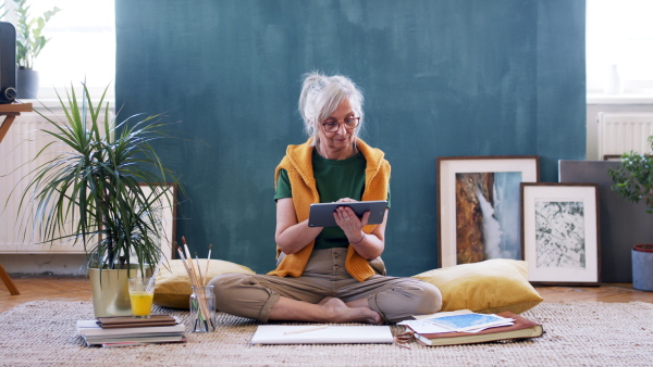 Portrait of senior woman designer with tablet sitting on floor at home, working.