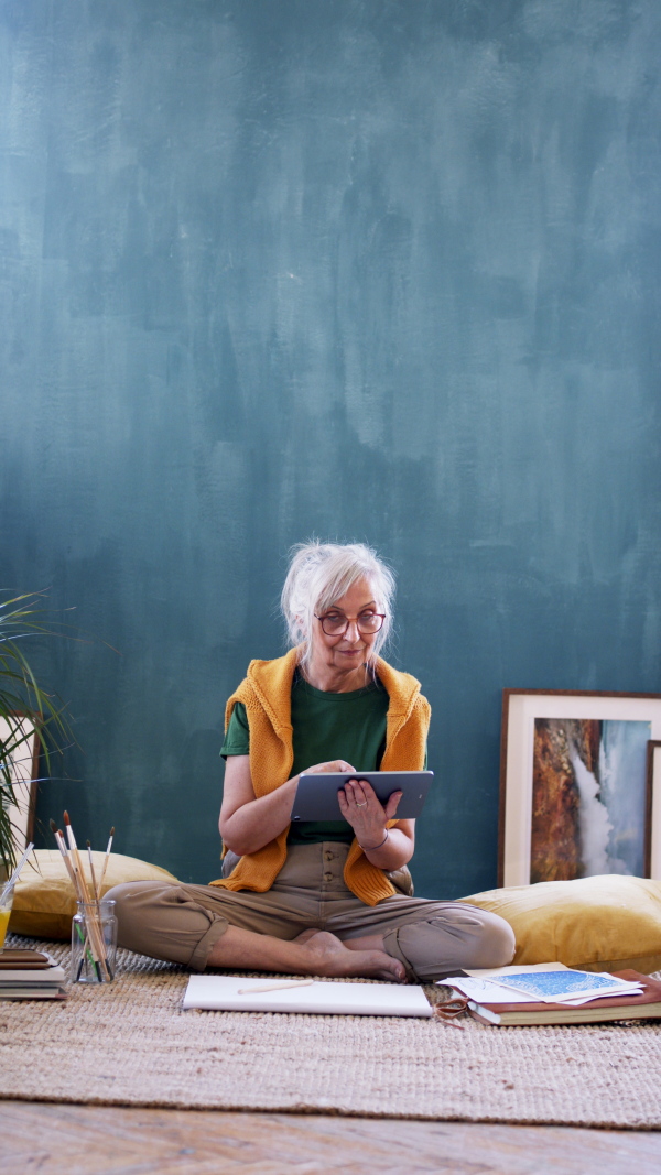 Portrait of senior woman designer with tablet sitting on floor at home, working, vertical video.