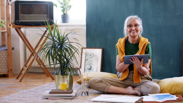 Portrait of senior woman designer with tablet sitting on floor at home, working.