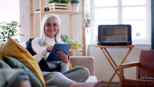 Portrait of senior woman sitting indoors on sofa at home, using tablet and looking at camera.