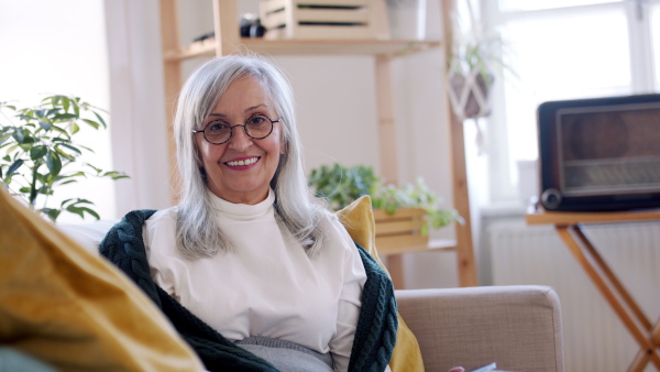 Portrait of senior woman sitting indoors on sofa at home, using tablet and looking at camera.