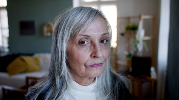 Front view portrait of sad senior woman standing indoors at home, looking at camera.