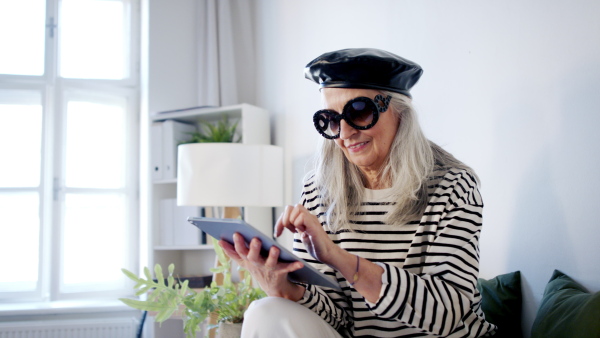 Portrait of senior woman sitting indoors on sofa at home, using tablet and looking at camera.