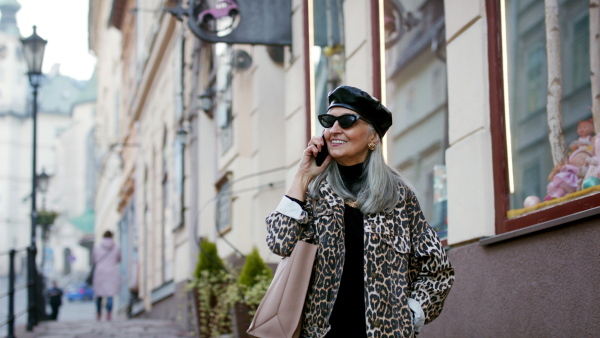 Senior cheerful chic woman with sunglasses standing outdoors, using smartphone.