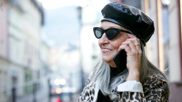 Senior cheerful chic woman with sunglasses standing outdoors, using smartphone.