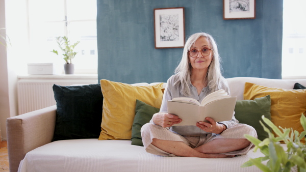 Portrait of senior woman sitting indoors on sofa at home, relaxing and reading book. Looking at camera.