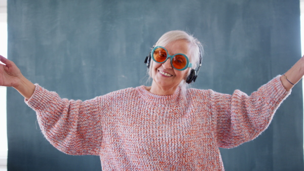 Portrait of senior woman with eccentric sunglasses standing indoors against dark background, looking at camera.