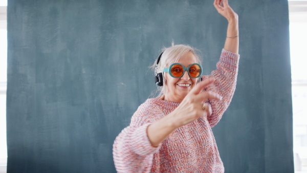 Portrait of senior woman with sunglases and headphones standing indoors against dark background, dancing.