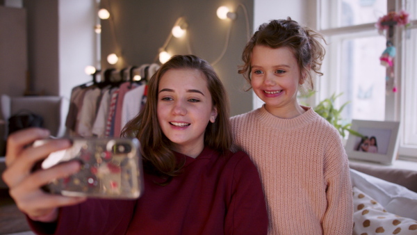 Portrait of sisters indoors at home, taking selfie with smartphone. Lockdown concept.