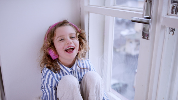 Small girl with headphones indoors at home, sitting on window sill, looking at camera.