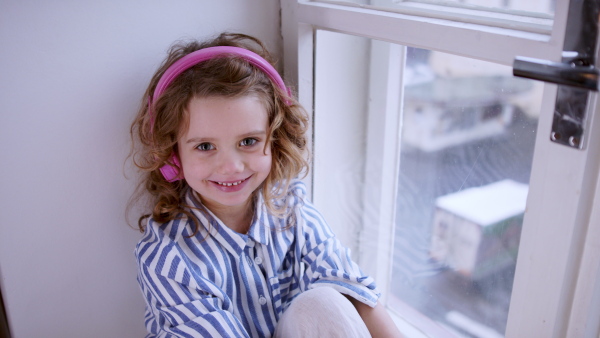 Portrait of sad small girl with headphones indoors at home, sitting on window sill.