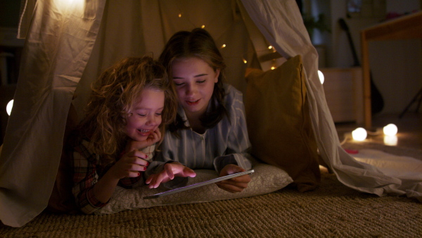 Sisters indoors at home, using tablet in pajamas in tent. Lockdown concept.