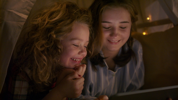 Sisters indoors at home, using tablet in pajamas in tent. Lockdown concept.