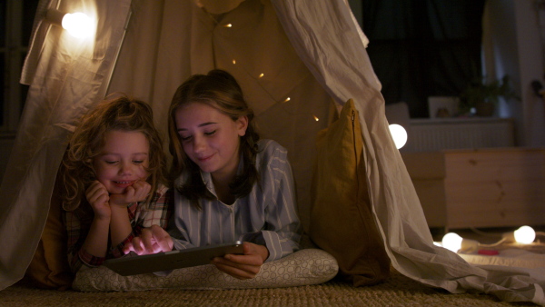 Sisters indoors at home, using tablet in pajamas in tent. Lockdown concept.