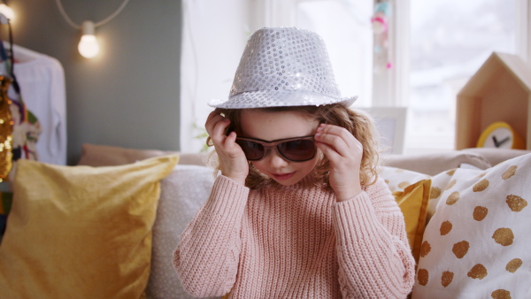 Portrait of small girl indoors at home, playing and dressing up on bed, looking at camera. Lockdown concept.