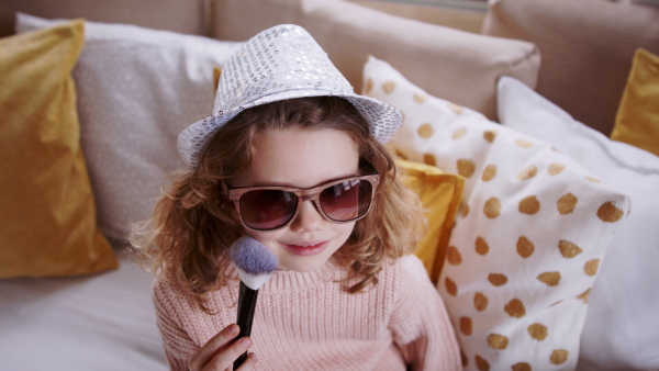 Front view of small girl with sunglasses indoors at home, playing with make-up brush on bed. Lockdown concept.