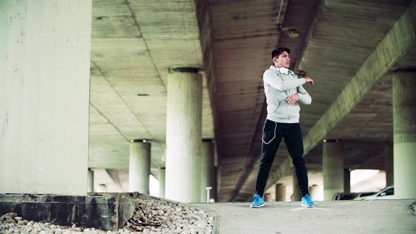 Young sporty man with headphones standing under the bridge in the city, stretching. Slow motion.