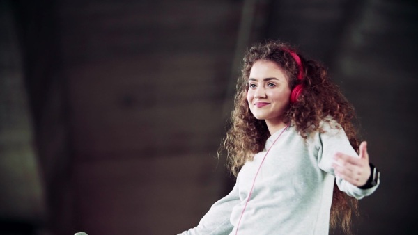 Beautiful young woman with smartphone and headphones standing under the bridge in the city, listening to music and dancing. Slow motion.