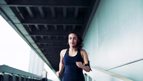 Beautiful young athlete with smartphone in an armband and earphones running in the city. Slow motion.