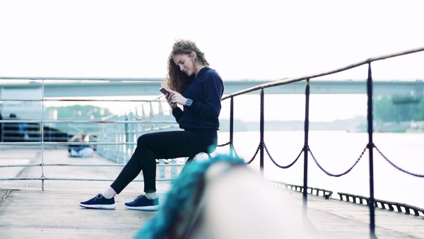 Sporty young woman with smartwatch sitting by the river in the city, using smartphone. Slow motion.