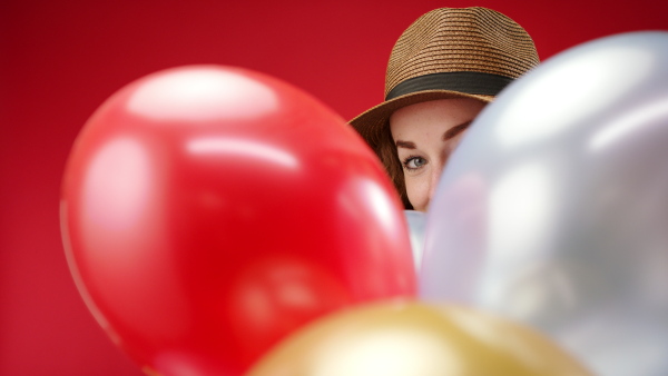 Young woman with hat and balloons in a studio on red background, looking at camera.