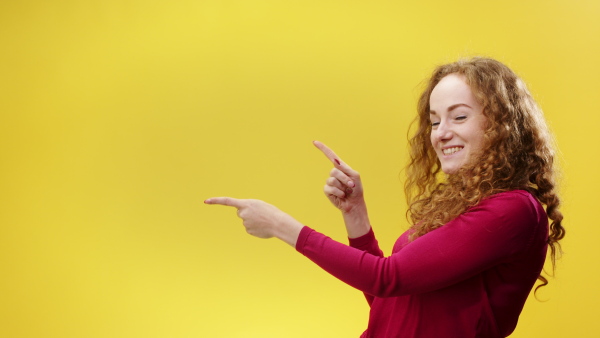 Portrait of young woman in a studio on yellow background, pointing fingers and looking at camera.