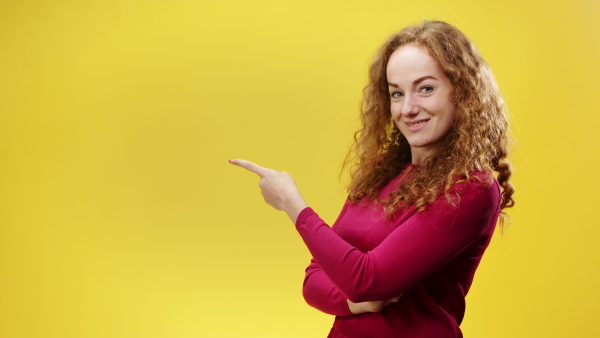 Portrait of young woman in a studio on yellow background, pointing finger and looking at camera.