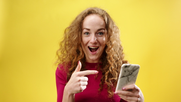 Surprised young woman in a studio on yellow background, pointing fingers to smartphone and looking at camera.