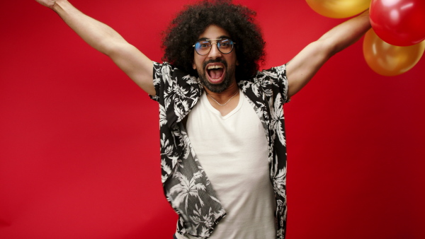 Young man celebrating with balloons in a studio on red background, looking at camera.