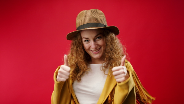 Young woman with hat in a studio on red background, looking at camera and making gestures.