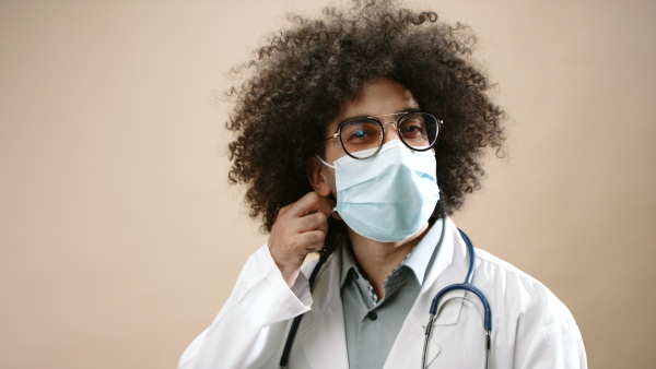 Mature man doctor with face mask standing in studio looking at camera, coronavirus concept. Copy space.