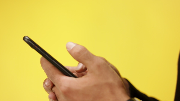 Close up of unrecognizable man using black smartphone in a studio on yellow background.