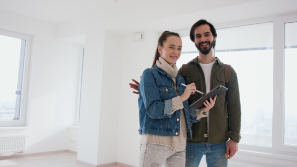 Happy young couple with tablet moving and planning in new flat, looking at camera. New home and relocation concept.