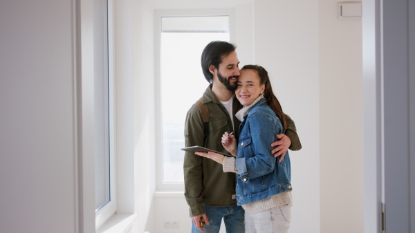 Happy young couple with tablet moving and planning in new flat, looking at camera. New home and relocation concept.