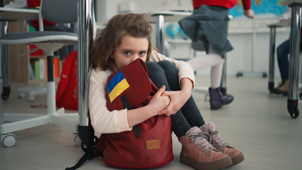 A little sad Ukrainian girl sitting on floor at school, concept of enrolling Ukrainian kids to schools.