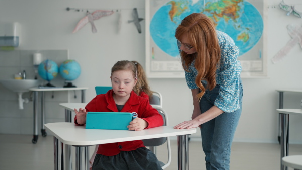 A Down syndrome schoolgirl using tablet with help of teacher during class at school, integration concept.