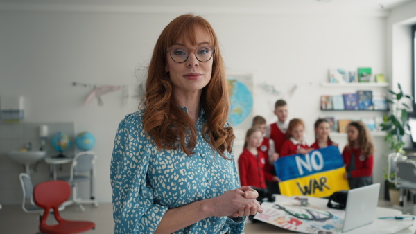 A young teacher looking at camera with little schoolchildren holidng and showing ukrainian flag in classroom at background, no war concept.