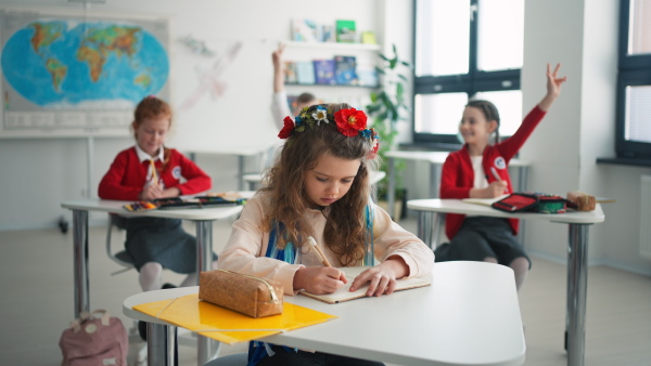 A little sad Ukrainian girl writing in classroom during class, concept of enrolling Ukrainian kids to schools.