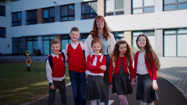 A portrait of happy schoolkids in uniforms with teacher at schoolyard, looking at camera.