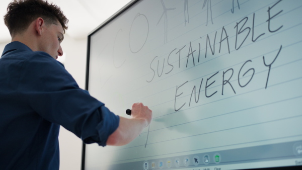 A young teacher writing on whiteboard during alternative energy lecture with group of senior students in classroom.